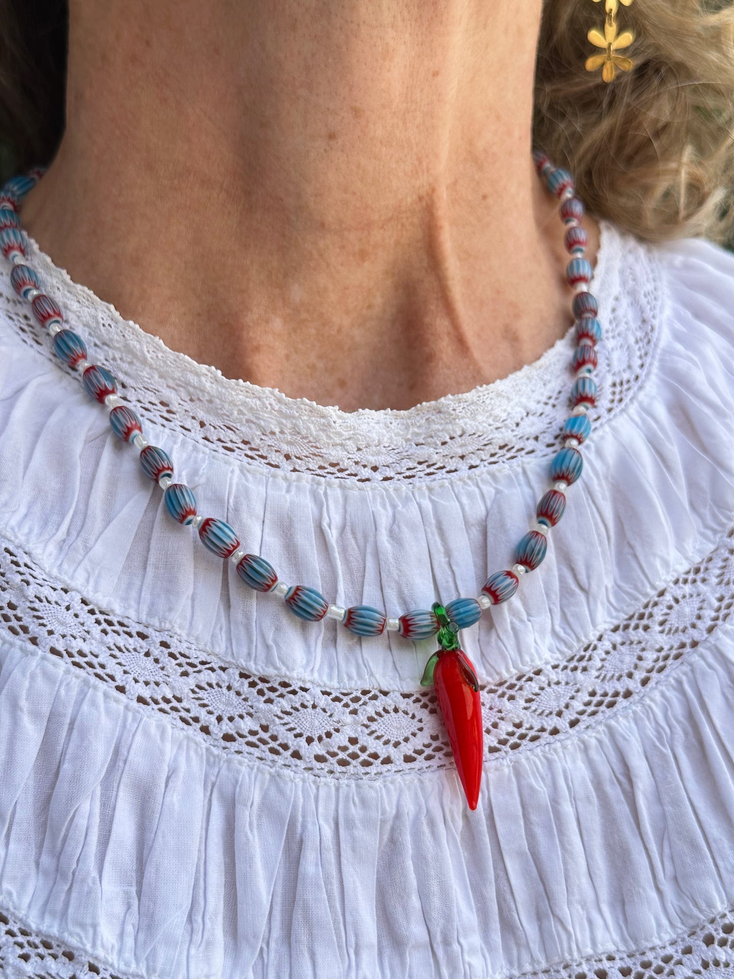 Collar hecho a mano con cuentas de cerámica azul y cuentas blancas con un abalorio con forma de chili de cristal de murano en color rojo y verde. Cierre ajustable de acero inoxidable.