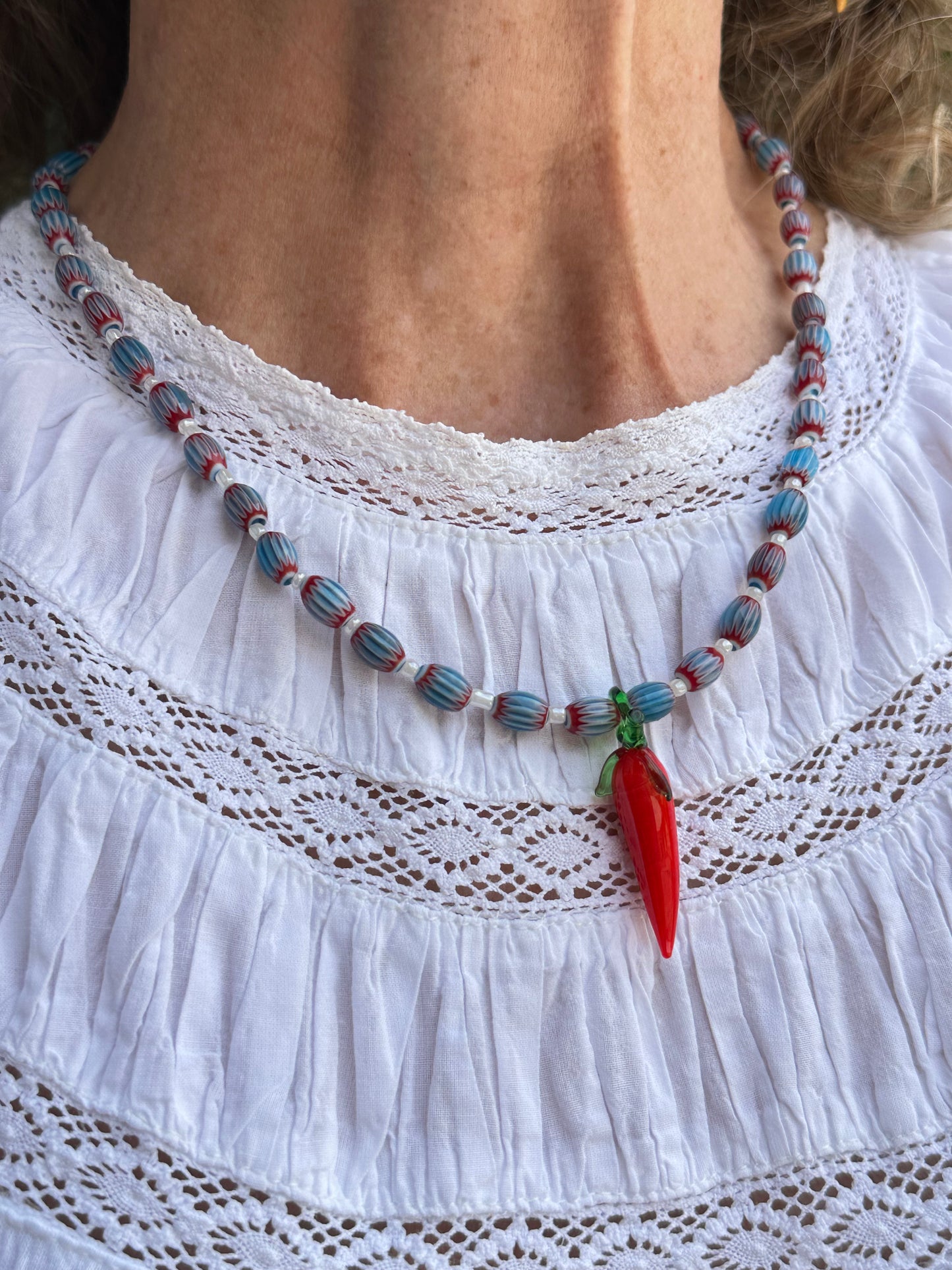 Collar hecho a mano con cuentas de cerámica azul y cuentas blancas con un abalorio con forma de chili de cristal de murano en color rojo y verde. Cierre ajustable de acero inoxidable.