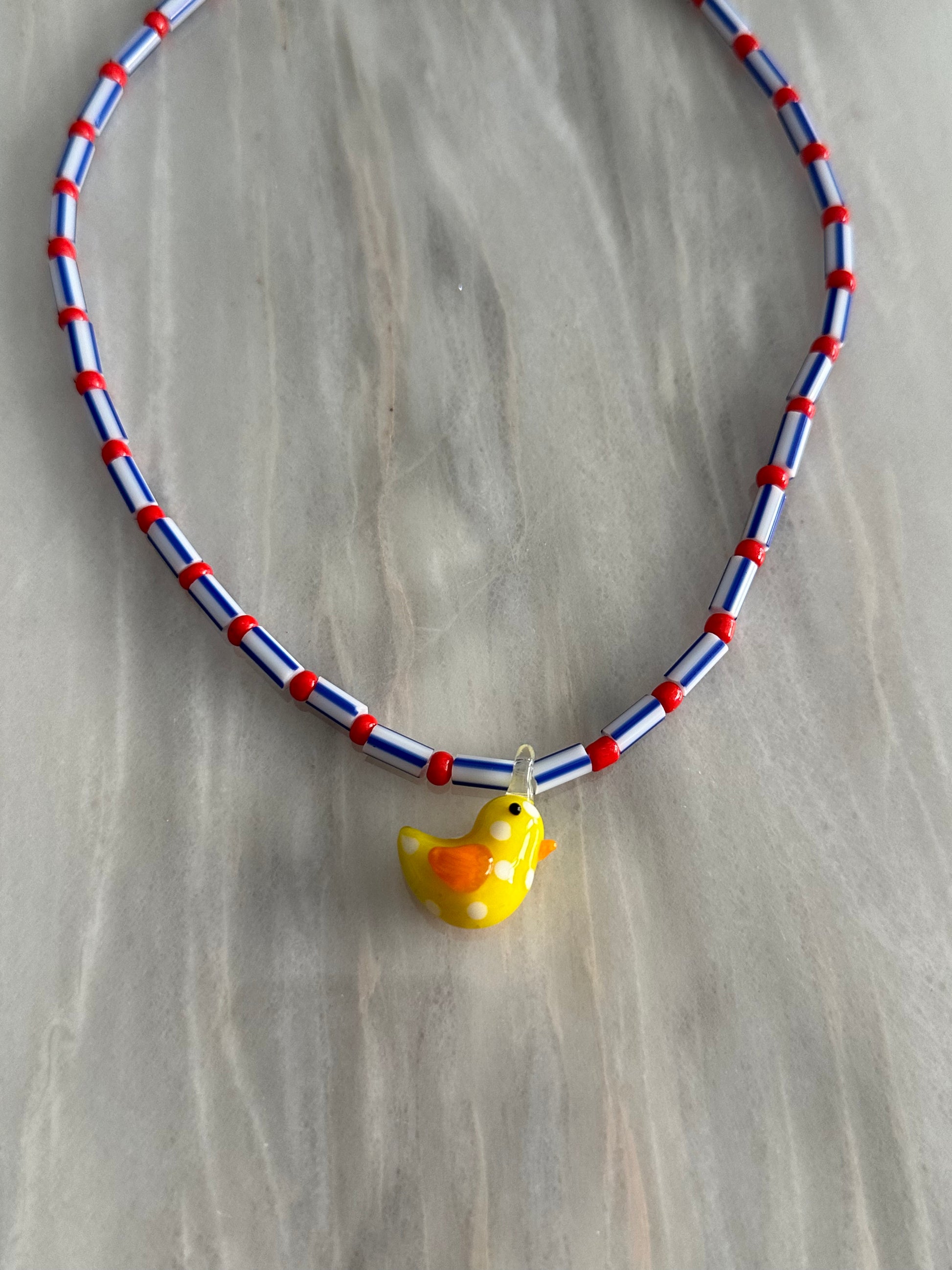 Collar hecho a mano con cuentas rayadas en azul, bolitas rojas y con un abalorio de cristal de murano con forma de pollito. Con cierre ajustable de acero inoxidable