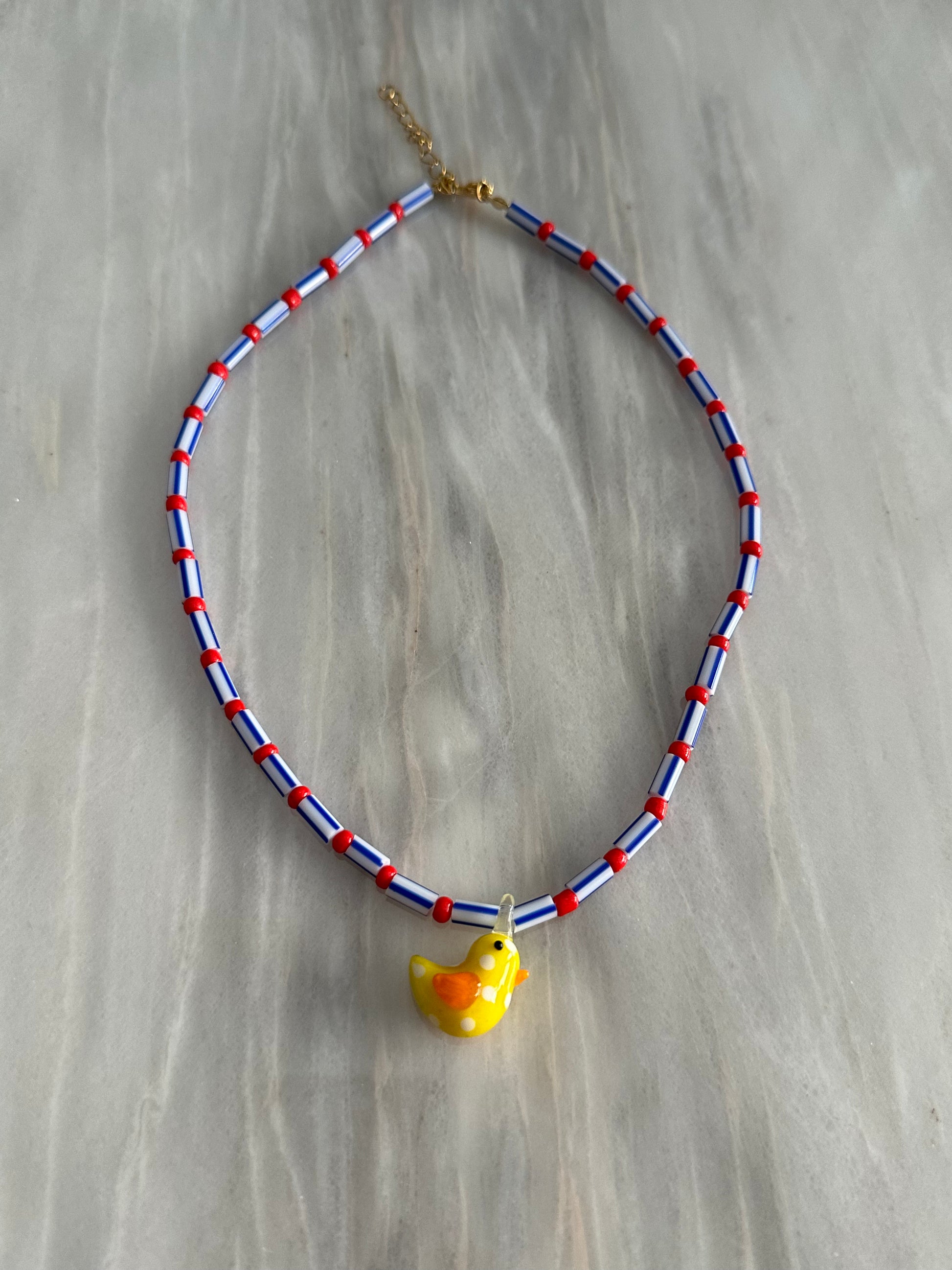 Collar hecho a mano con cuentas rayadas en azul, bolitas rojas y con un abalorio de cristal de murano con forma de pollito. Con cierre ajustable de acero inoxidable