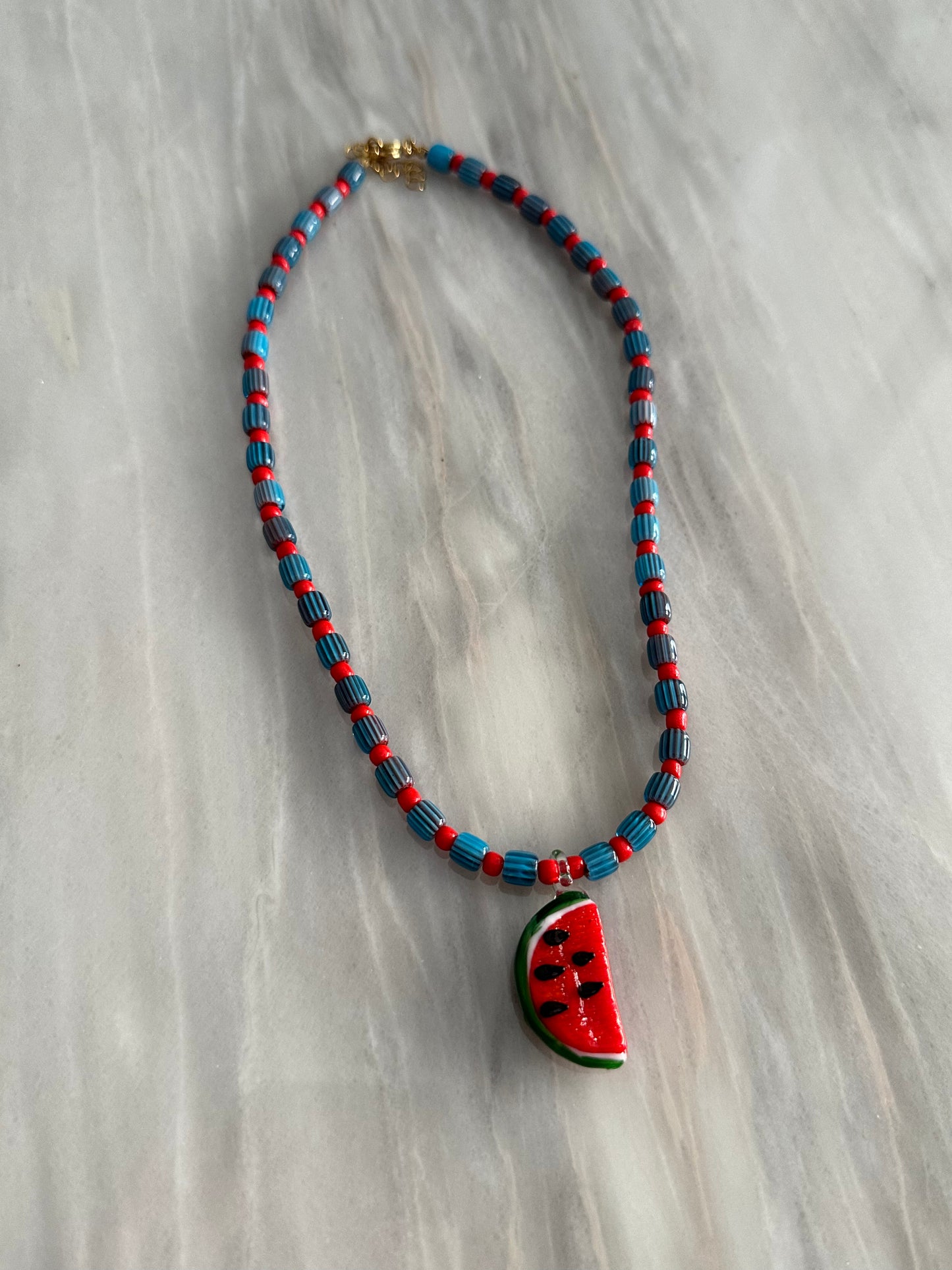 Collar hecho a mano con cuentas de cerámica azul y bolitas rojas, con un abalorio con forma de sandía de cristal de murano en color rojo y verde. Cierre ajustable de acero inoxidable