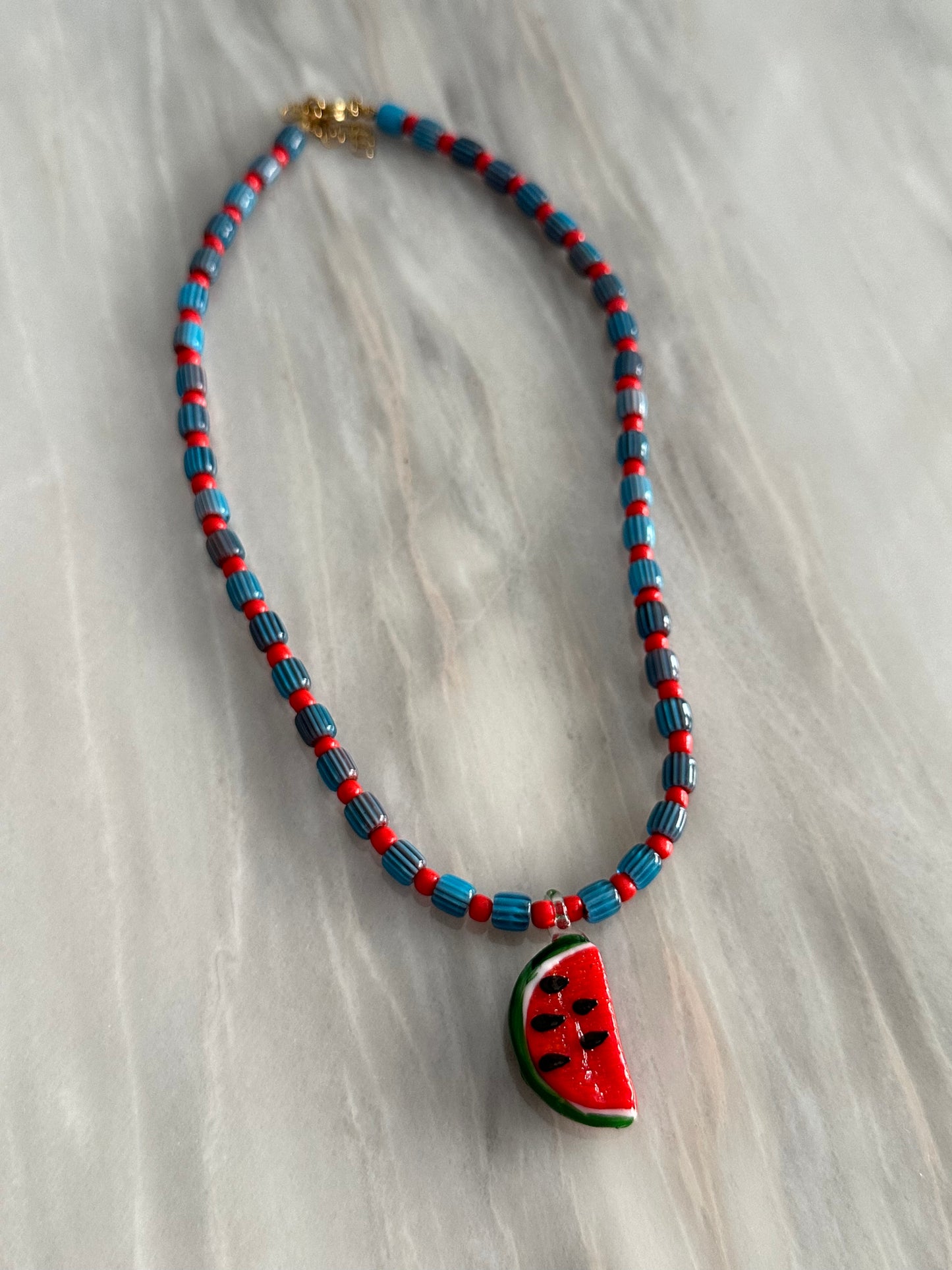 Collar hecho a mano con cuentas de cerámica azul y bolitas rojas, con un abalorio con forma de sandía de cristal de murano en color rojo y verde. Cierre ajustable de acero inoxidable