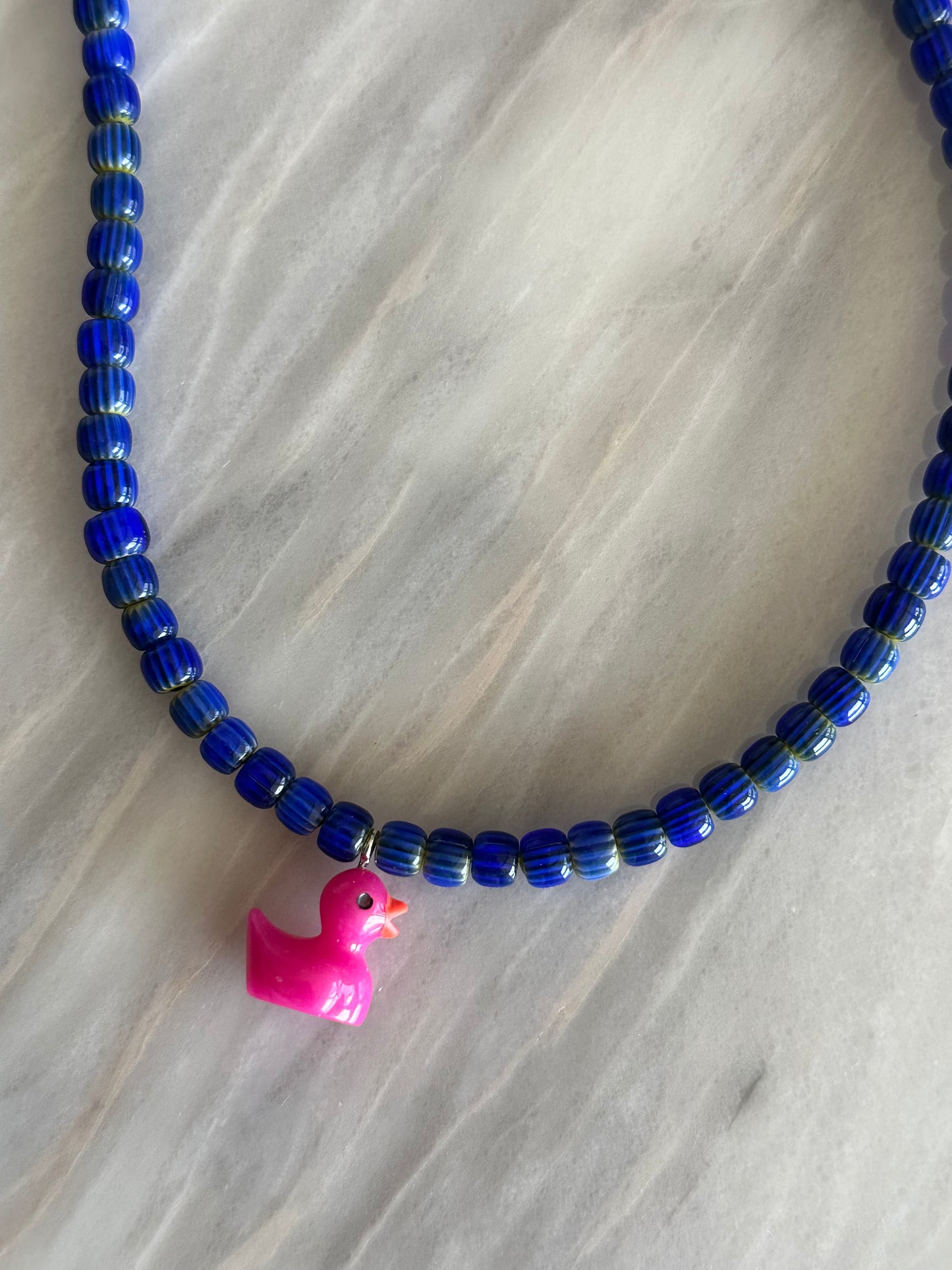 Collar hecho a mano con cuentas de cerámica azul y con un abalorio con forma de patito en color rosa. Cierre ajustable de acero inoxidable