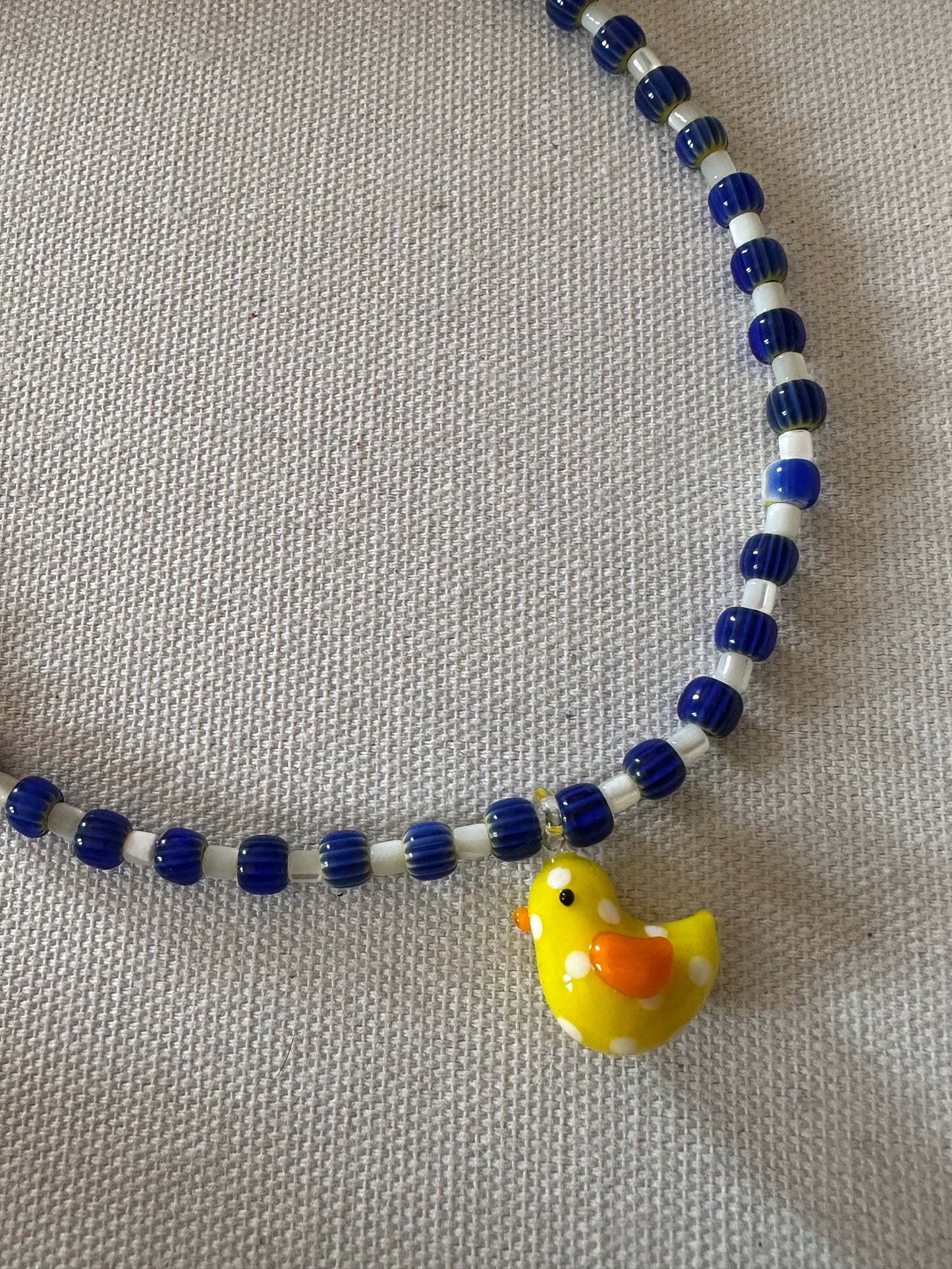 Collar hecho a mano con cerámica azul marino, cuentas blancas y con un abalorio de cristal de murano con forma de pollito en color amarillo. Con cierre ajustable de acero inoxidable