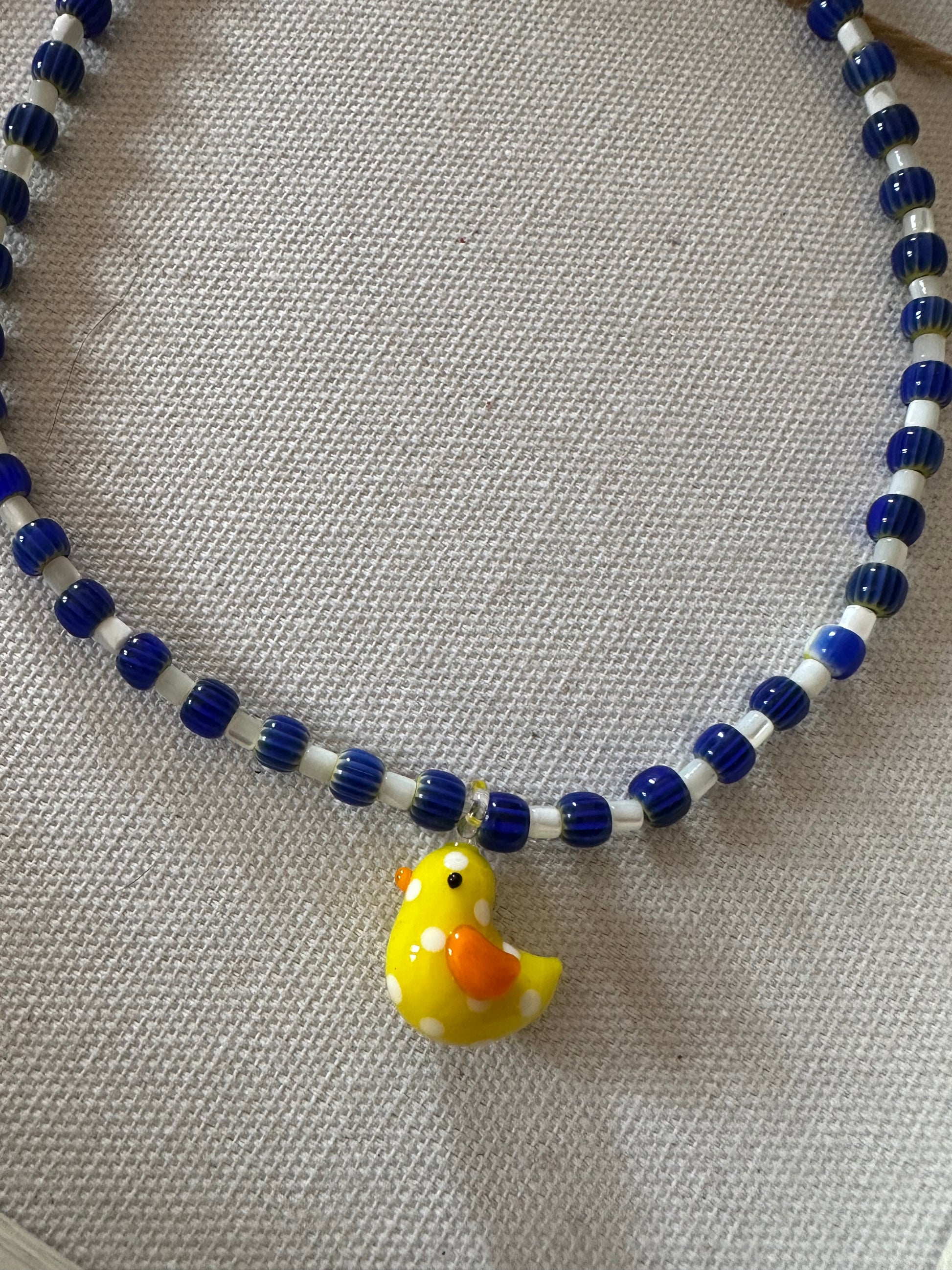 Collar hecho a mano con cerámica azul marino, cuentas blancas y con un abalorio de cristal de murano con forma de pollito en color amarillo. Con cierre ajustable de acero inoxidable