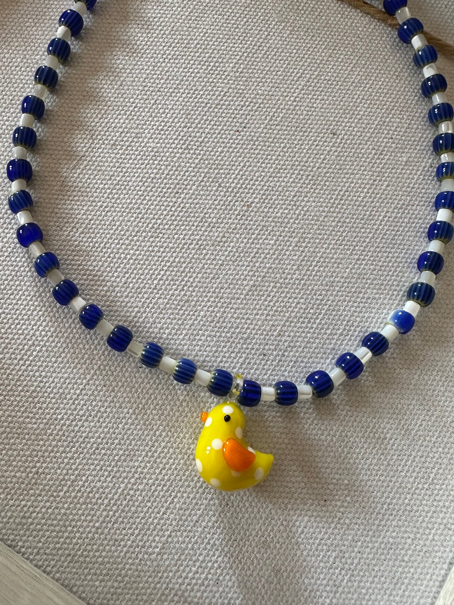 Collar hecho a mano con cerámica azul, cuentas blancas y con un abalorio de cristal de murano con forma de pollito en color amarillo. Con cierre ajustable de acero inoxidable