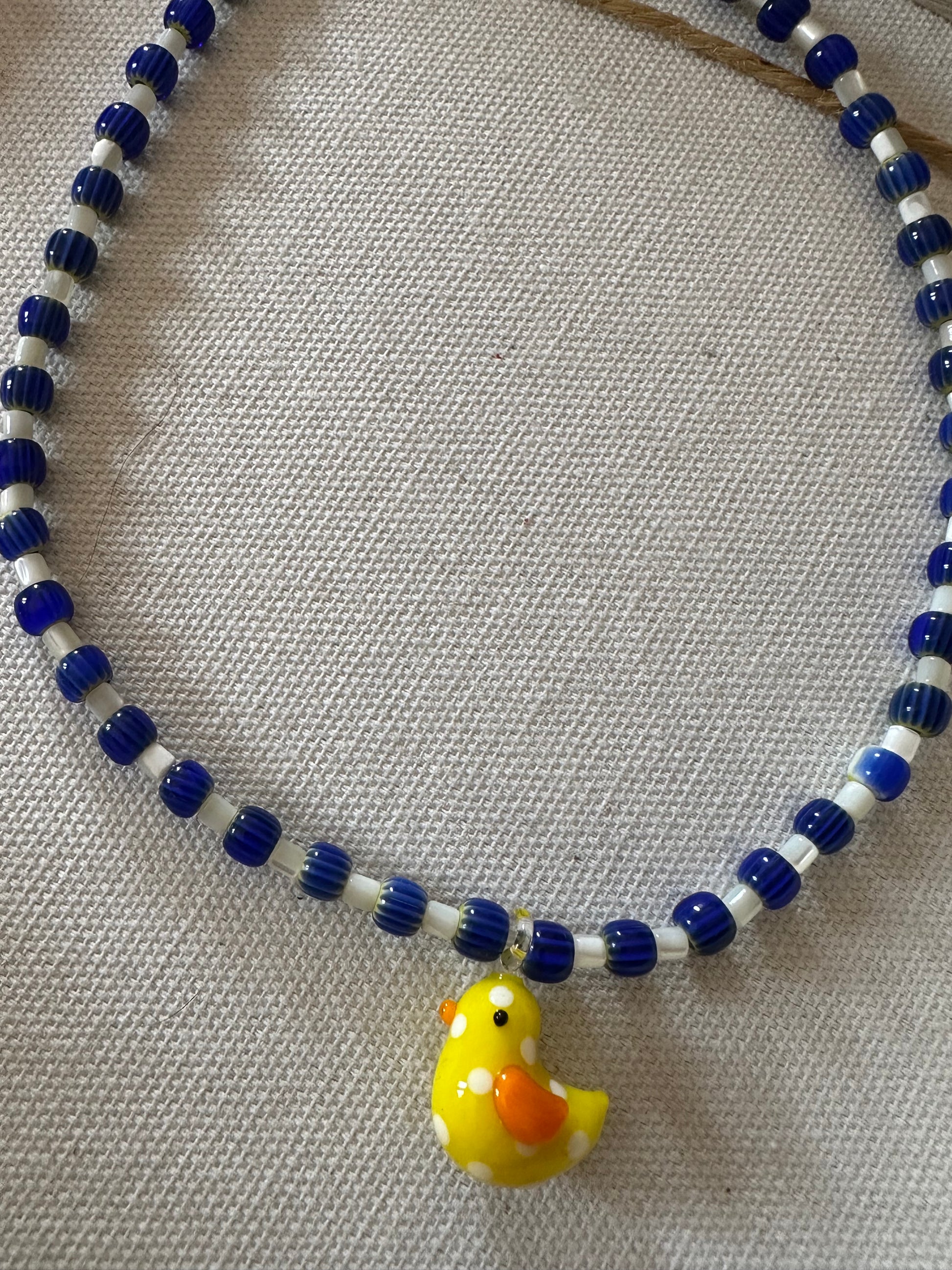 Collar hecho a mano con cerámica azul marino, cuentas blancas y con un abalorio de cristal de murano con forma de pollito en color amarillo. Con cierre ajustable de acero inoxidable