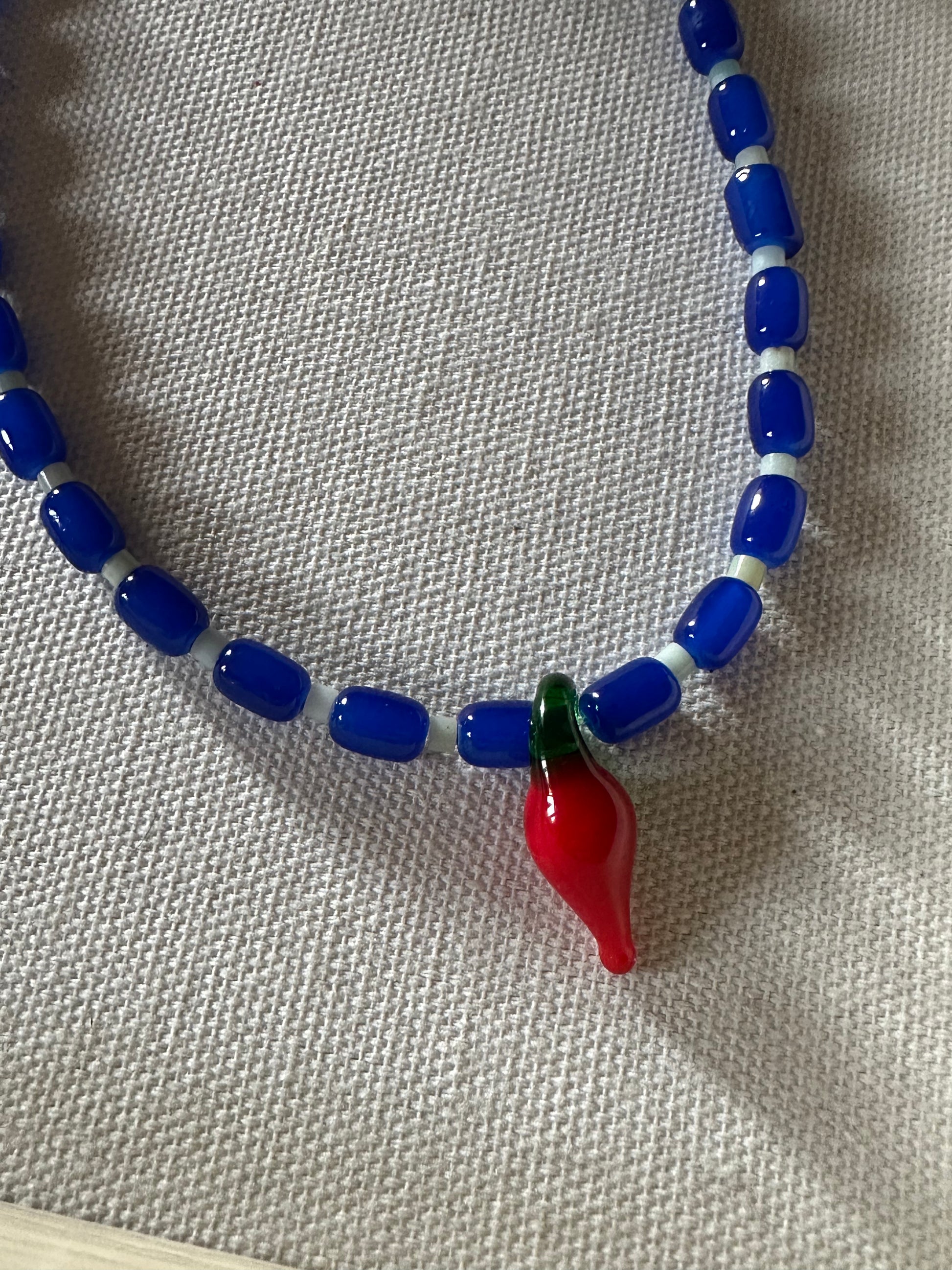 Collar hecho a mano con cuentas de cerámica azul y bolitas blancas, con un abalorio con forma de chili de cristal de murano en color rojo y verde. Cierre ajustable