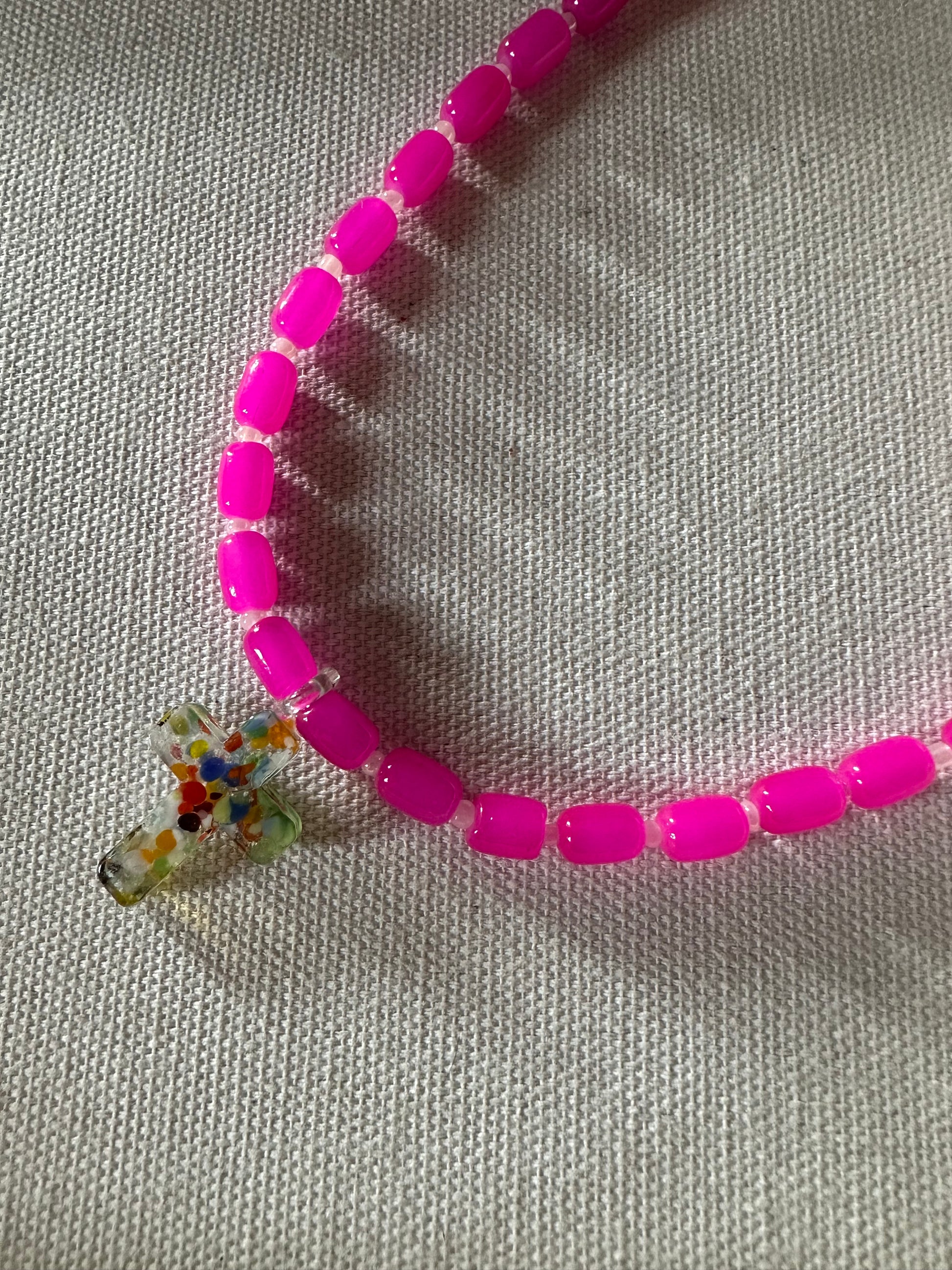 Collar hecho a mano con cuentas de cerámica rosa y con un abalorio con forma de cruz de cristal de murano en varios tonos. Cierre ajustable de acero inoxidable