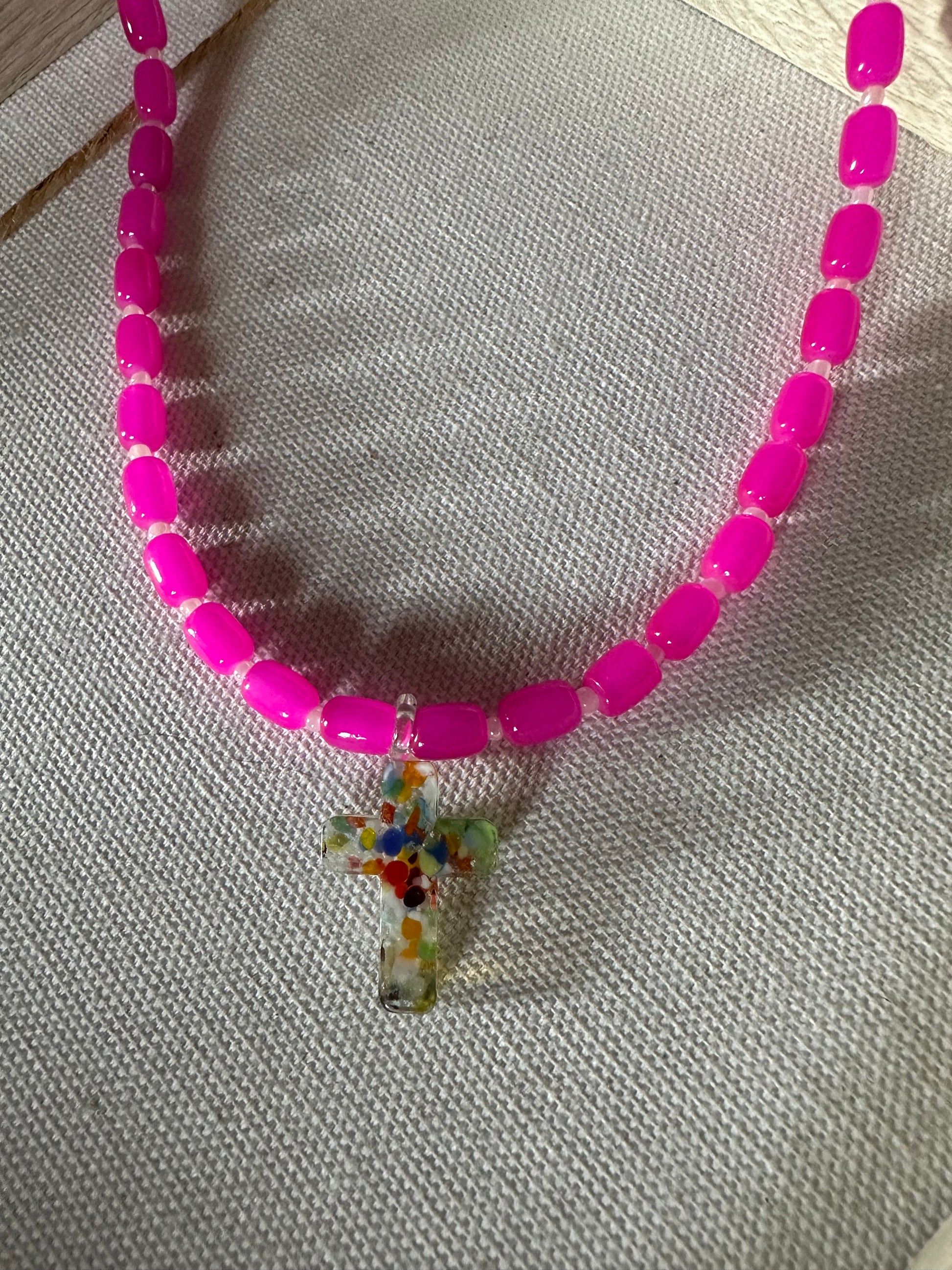 Collar hecho a mano con cuentas de cerámica rosa y con un abalorio con forma de cruz de cristal de murano en varios tonos. Cierre ajustable de acero inoxidable